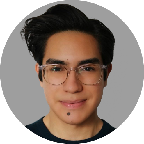 A close-up photo of a young man
										with a warm complexion, sporting sleek,
										dark hair styled upward. They are
										wearing clear-framed glasses and a
										dark, casual top. The person has a
										subtle smile and a beauty mark just
										below the lower lip. The background is
										simple grey color.