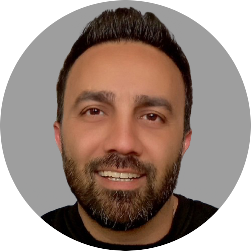 A close-up portrait of a man with
										an olive complexion, dark eyes, and a
										full beard. They have short, dark hair
										that is styled back and are smiling
										with a closed mouth. The individual is
										wearing a black top and the background
										is a simple grey color.